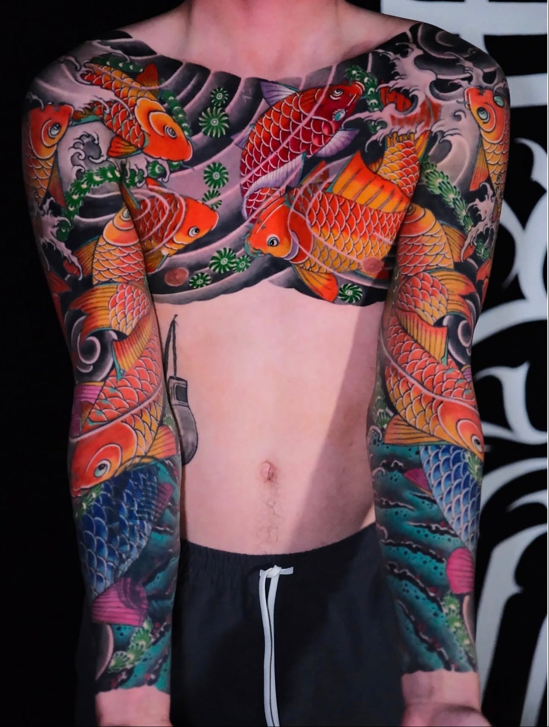 Person with colorful koi fish tattoos on arms and chest against a black and white patterned background